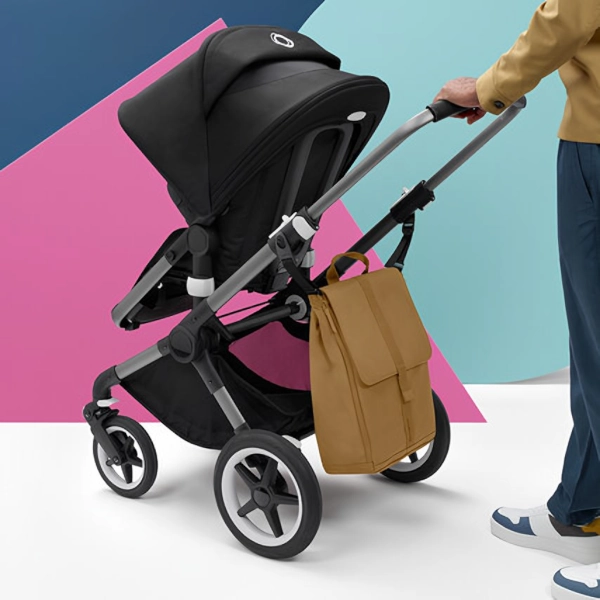 Ein Kinderwagen mit schwarzem Sitz und einer beigefarbenen Bugaboo Wickelrucksack, die seitlich am Griff befestigt ist, vor einem bunten Hintergrund.