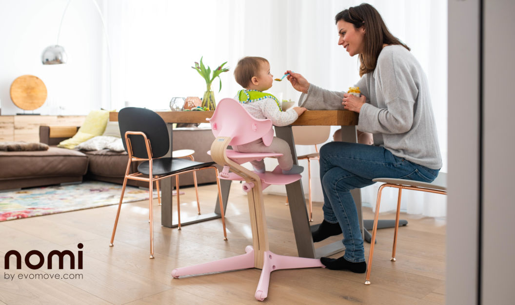Mutter füttert Ihr Kind, welches in einem Nomi Hochstuhl mit Mini Bügel sitzt
