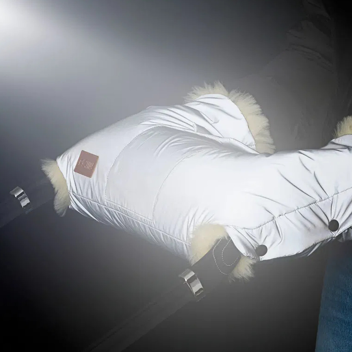 Silberner Handwärmer mit reflektierender Oberfläche am Griff eines Kinderwagens, beleuchtet von einer Lichtquelle im Dunkeln. Das Lammfellfutter schaut an den Rändern hervor.