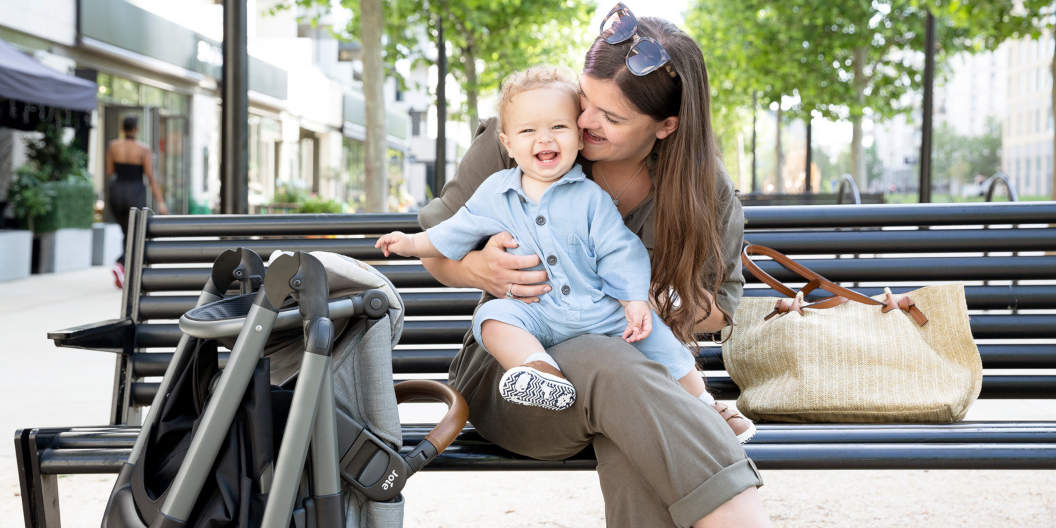 Mutter sitzt mit Kind auf einer Parkbank mit gefaltetem Joie Aeria Kinderwagen bei sich
