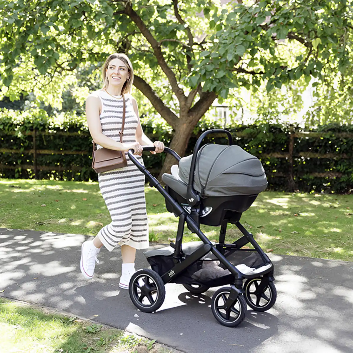 Eine Frau spaziert fröhlich mit dem Kinderwagen und Babyschale durch einen sonnigen Park.