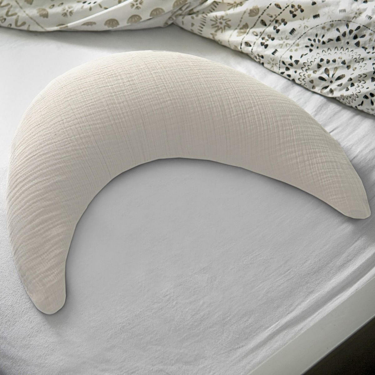 Ein Theraline Musselinmond liegt auf einem Bett mit weißem Spannbettlaken und gemusterter Bettdecke.