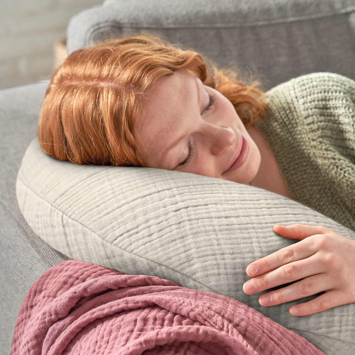 Eine Frau mit roten Haaren sitzt lächelnd auf dem Sofa, hält ein Tablet in der Hand und hat das Theraline Musselinmond bequem auf dem Schoß.